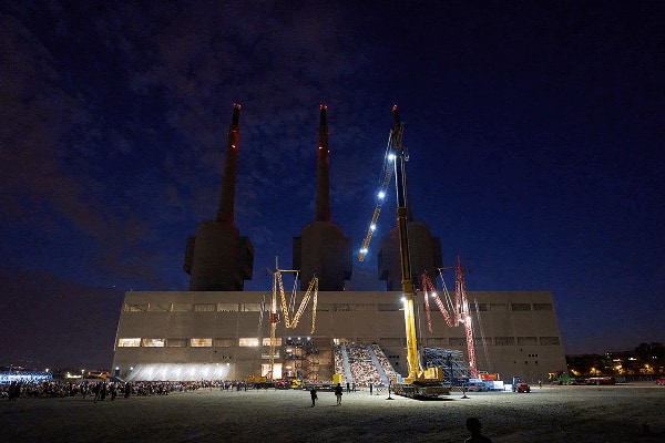Three Chimneys power plant illuminated at night with cranes positioned in front of the building.